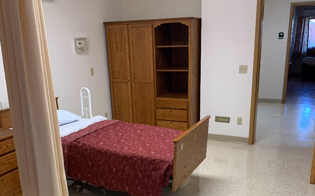 Small resident bedroom with a single bed covered by a red blanket, wooden wardrobe and shelving, and an open doorway to a corridor.
