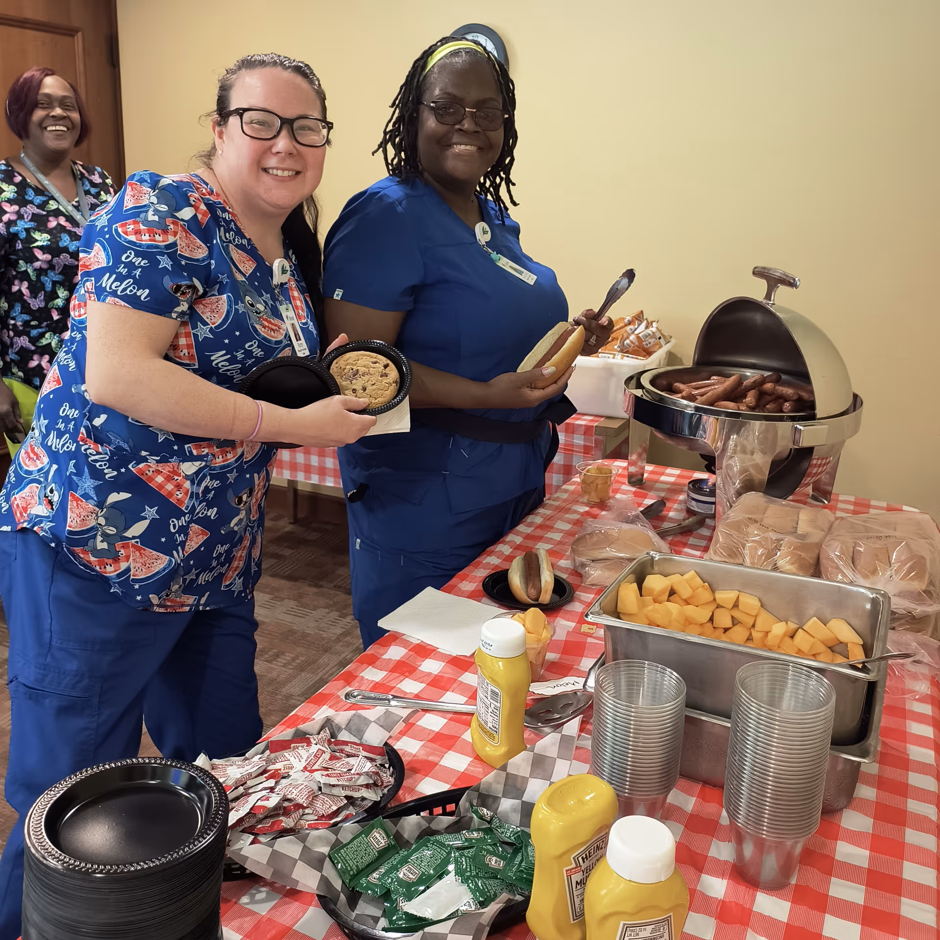 Three people in scrubs smiling as they serve hot dogs and sides from a buffet table with a red checkered tablecloth.