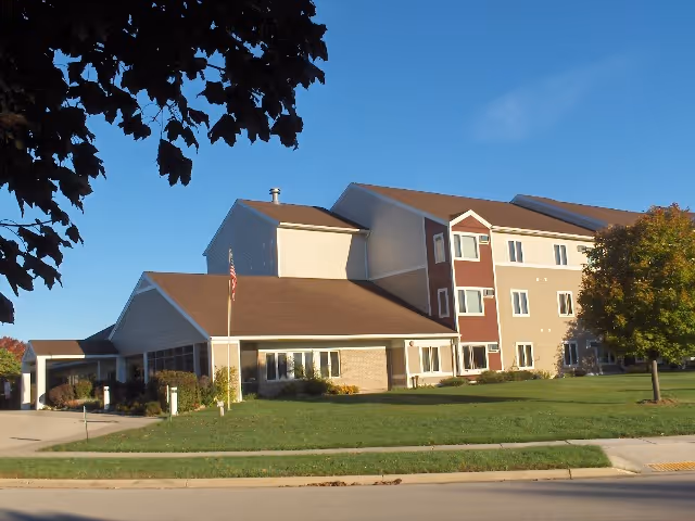 Exterior view of a multi-story senior living facility building with beige and brown walls, a sloped roof, and several windows. There is a well-maintained lawn with a tree and an American flag near the entrance. The sky is clear and blue.