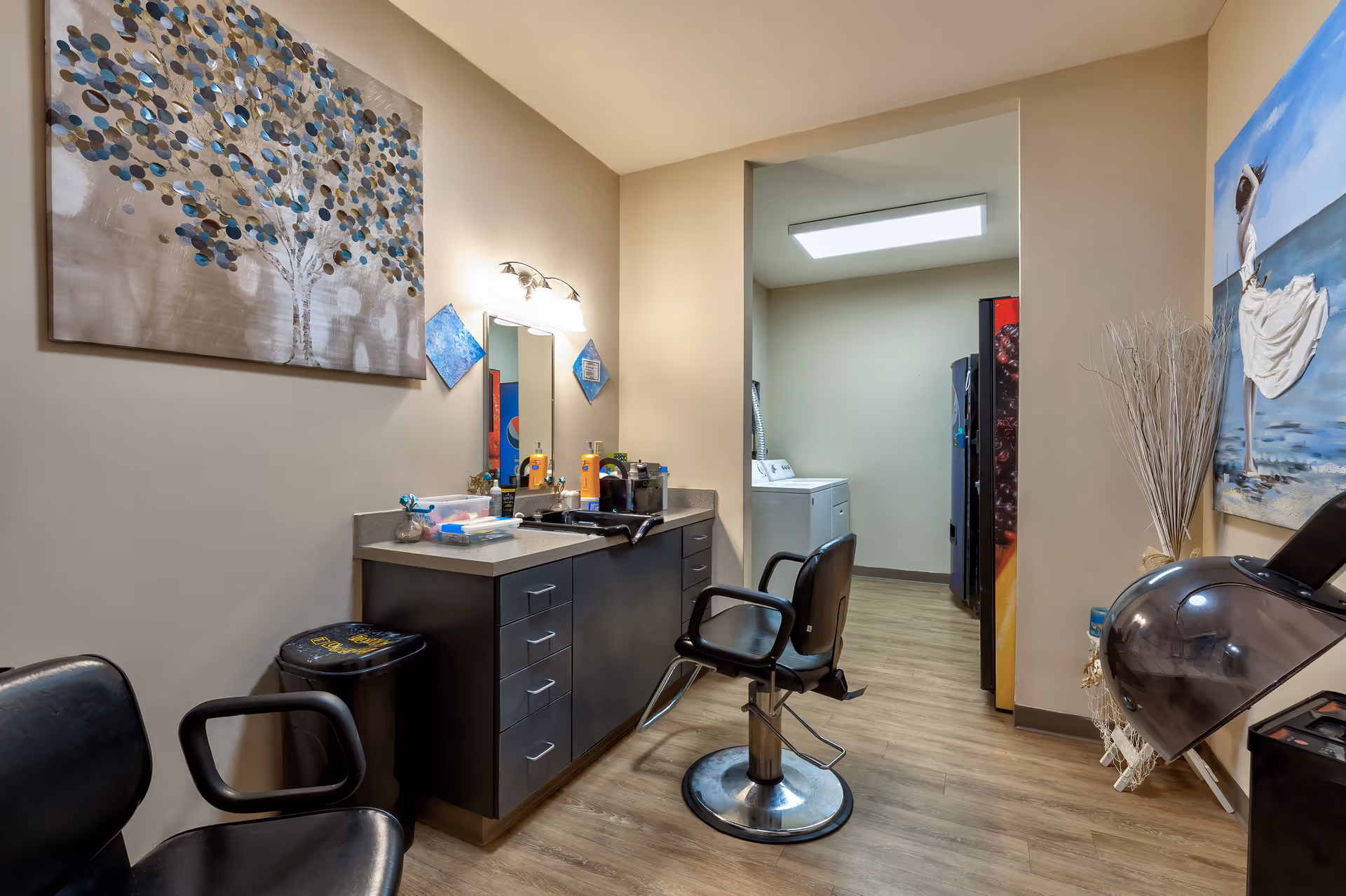 Interior view of a salon area in a facility with two black salon chairs, a countertop with various hair products and tools, a mirror with lights above it, and decorative paintings on the walls. In the background, there is a laundry area with a washing machine and a refrigerator.