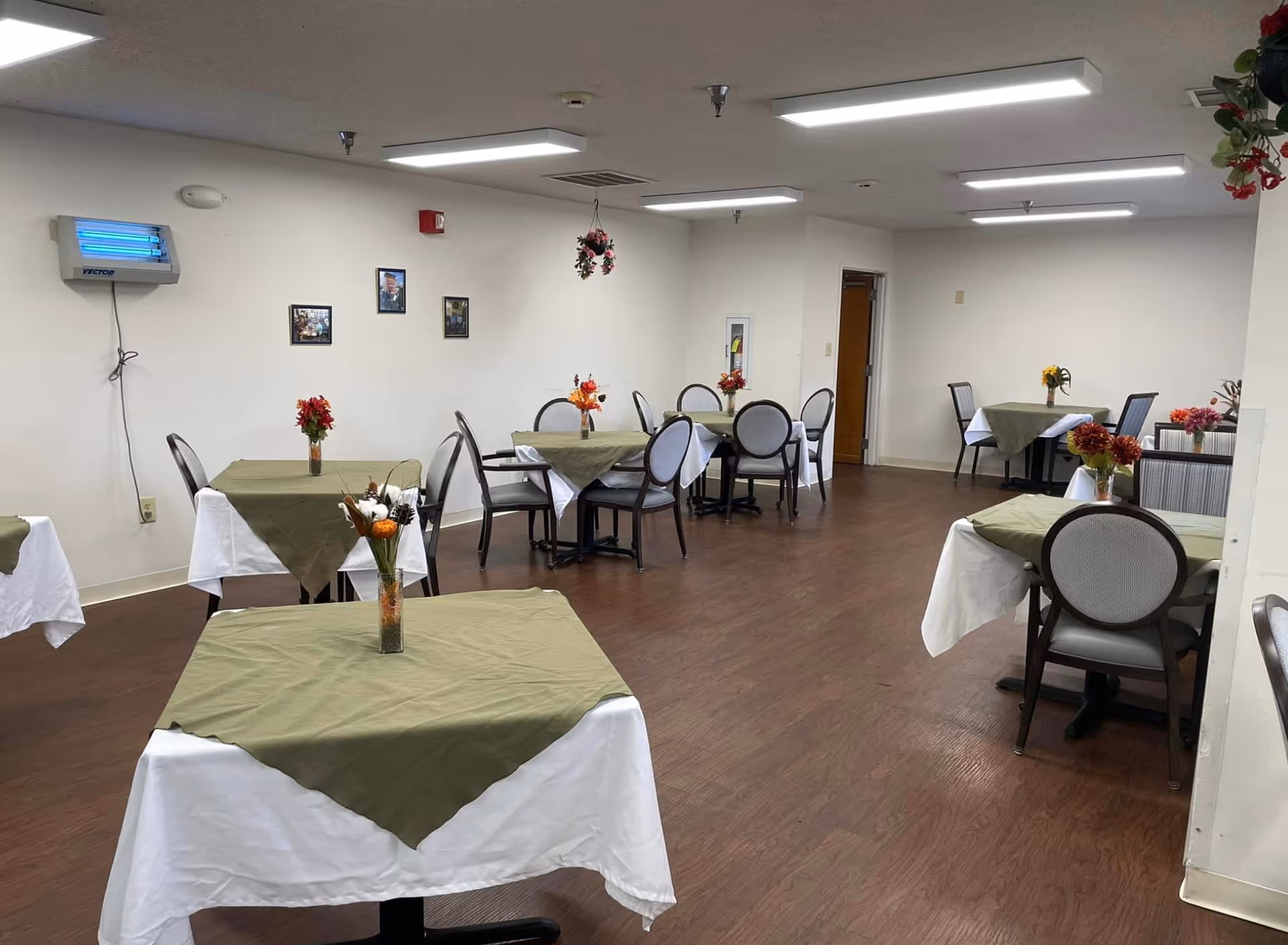 A dining room with several tables covered with white tablecloths and green overlays, each table decorated with a small vase of flowers. The room has wooden flooring, white walls with a few framed pictures, and bright overhead lighting. Chairs with cushioned seats and backs are arranged around the tables.