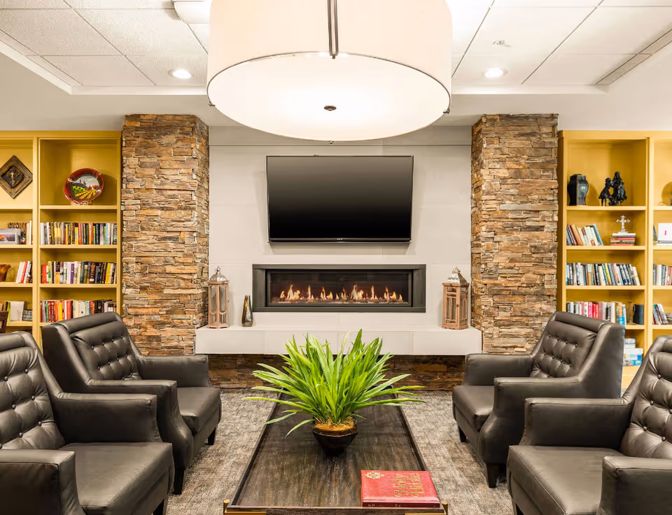 Cozy common room with a central coffee table and potted plant, surrounded by leather armchairs, a fireplace and wall-mounted TV flanked by stone columns and bookshelves.