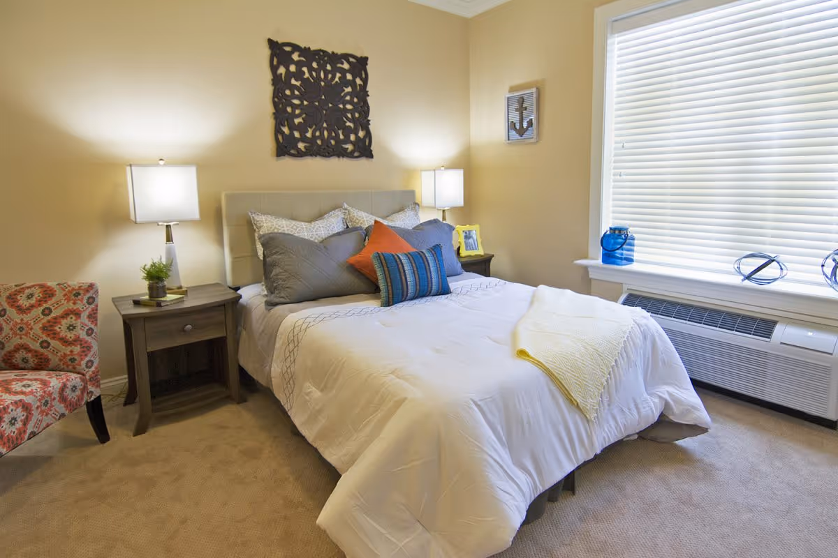 A cozy bedroom with a neatly made bed featuring multiple pillows in gray, orange, and blue colors. There are two bedside tables with lamps on each side of the bed. A decorative wall piece hangs above the bed, and a window with closed blinds is on the right side. A patterned armchair is partially visible on the left side of the image.