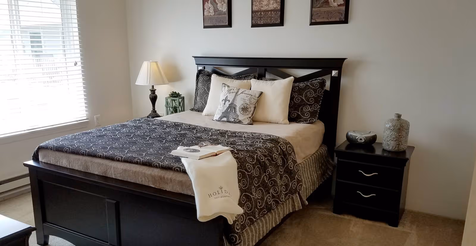 A neatly made bed with a black wooden frame and patterned black and beige bedding in a bedroom. There are five pillows on the bed, including one with an Eiffel Tower design. A white towel with the word 'Holiday' embroidered on it is draped over the foot of the bed. To the right of the bed is a black nightstand with two drawers, holding a decorative vase and a small radio. A lamp and a green lantern are on the left side of the bed near a window with white blinds. Three framed artworks hang on the wall above the bed.
