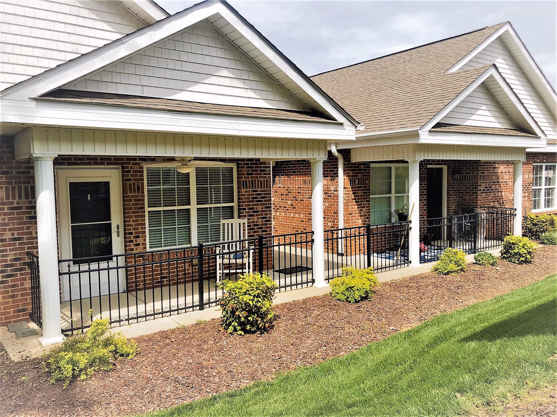 Exterior view of a brick building with white columns and a covered porch area featuring black metal railings. There are two doors and multiple windows with white shutters, along with small bushes and a well-maintained lawn in front.