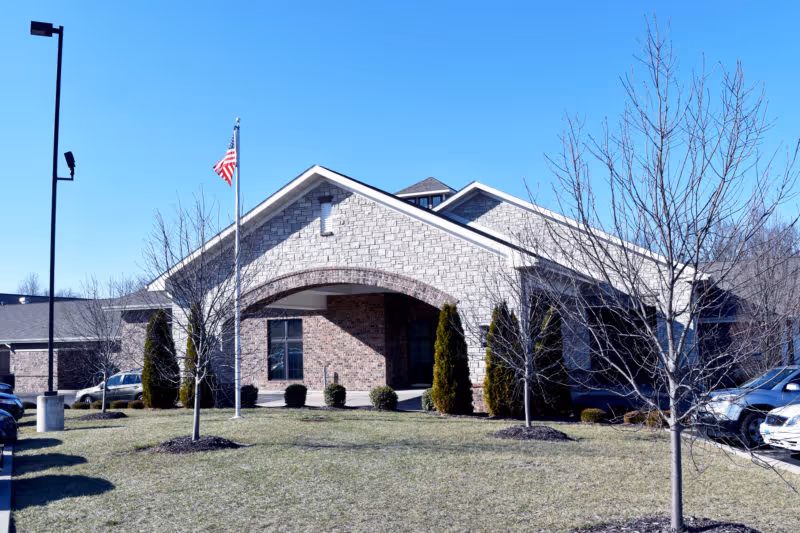 Front exterior view of Cedarhurst Senior Living of Waterloo building with a brick facade, an American flag on a flagpole, leafless trees, and parked cars on either side under a clear blue sky.