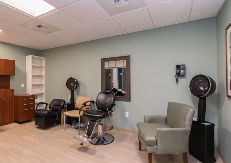 A room with light green walls and light wood flooring featuring salon-style chairs including a black barber chair, a black salon chair, and a beige chair. There are two black hair dryer stations, a wall-mounted telephone, a framed mirror on the wall, a green upholstered armchair, and wooden cabinets and shelving on the left side.