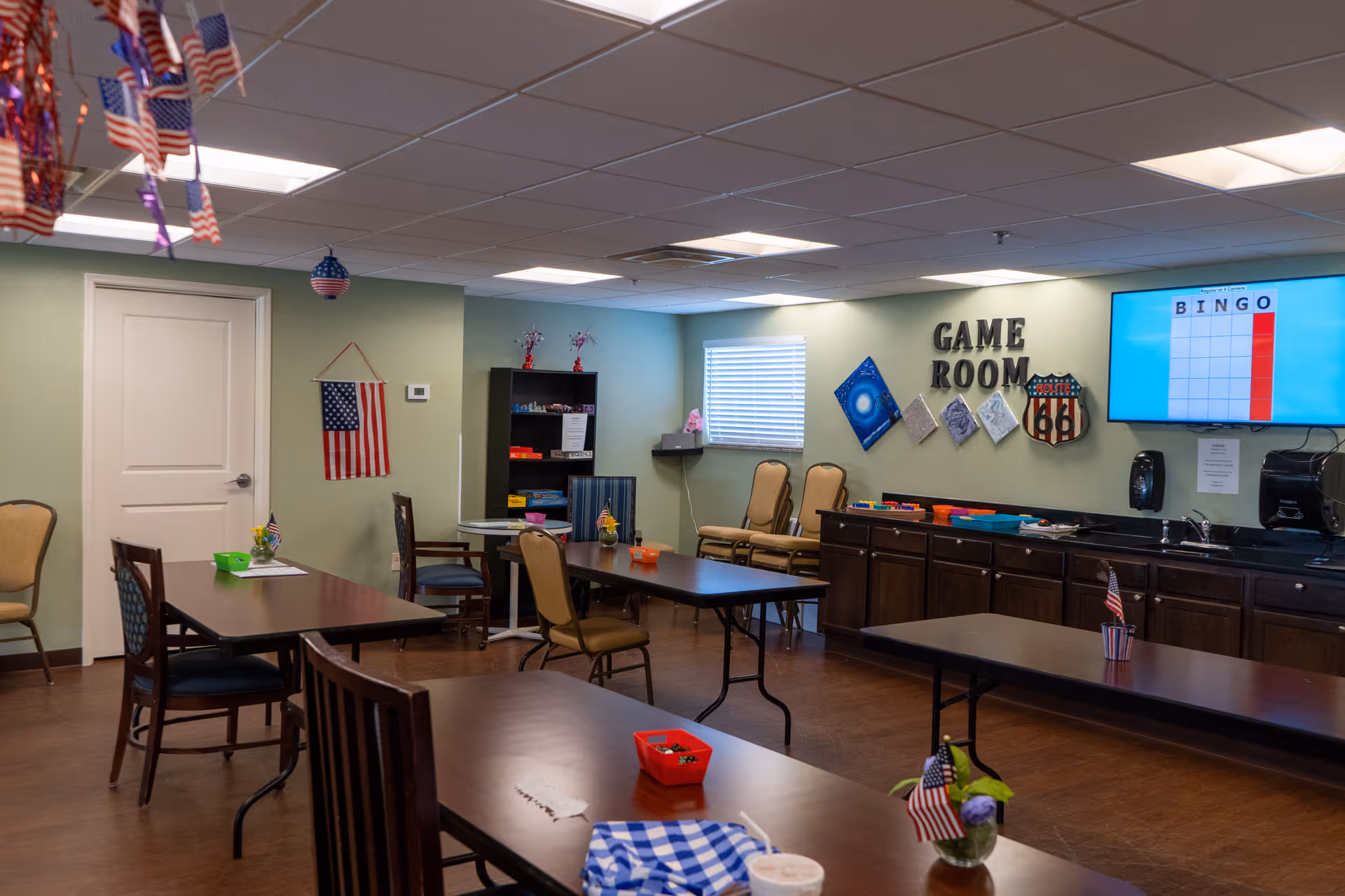 Interior view of a game room with several tables and chairs arranged around the room. The walls are light green with a window covered by blinds. On one wall, there is a sign that reads 'GAME ROOM' along with decorative items and a Route 66 sign. A large TV screen displays a bingo game. The room has a cabinet with a countertop and various items on it. Small American flags and patriotic decorations are placed on the tables and hanging from the ceiling.