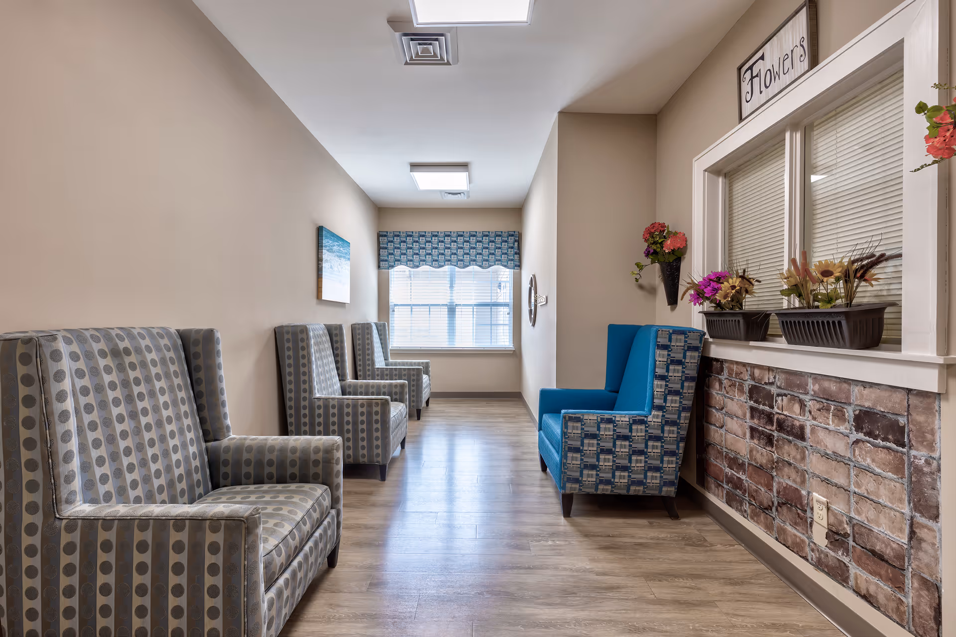 Bright interior hallway seating area with patterned armchairs, a window at the end, and decorative flowers under a sign reading 'Flowers'.