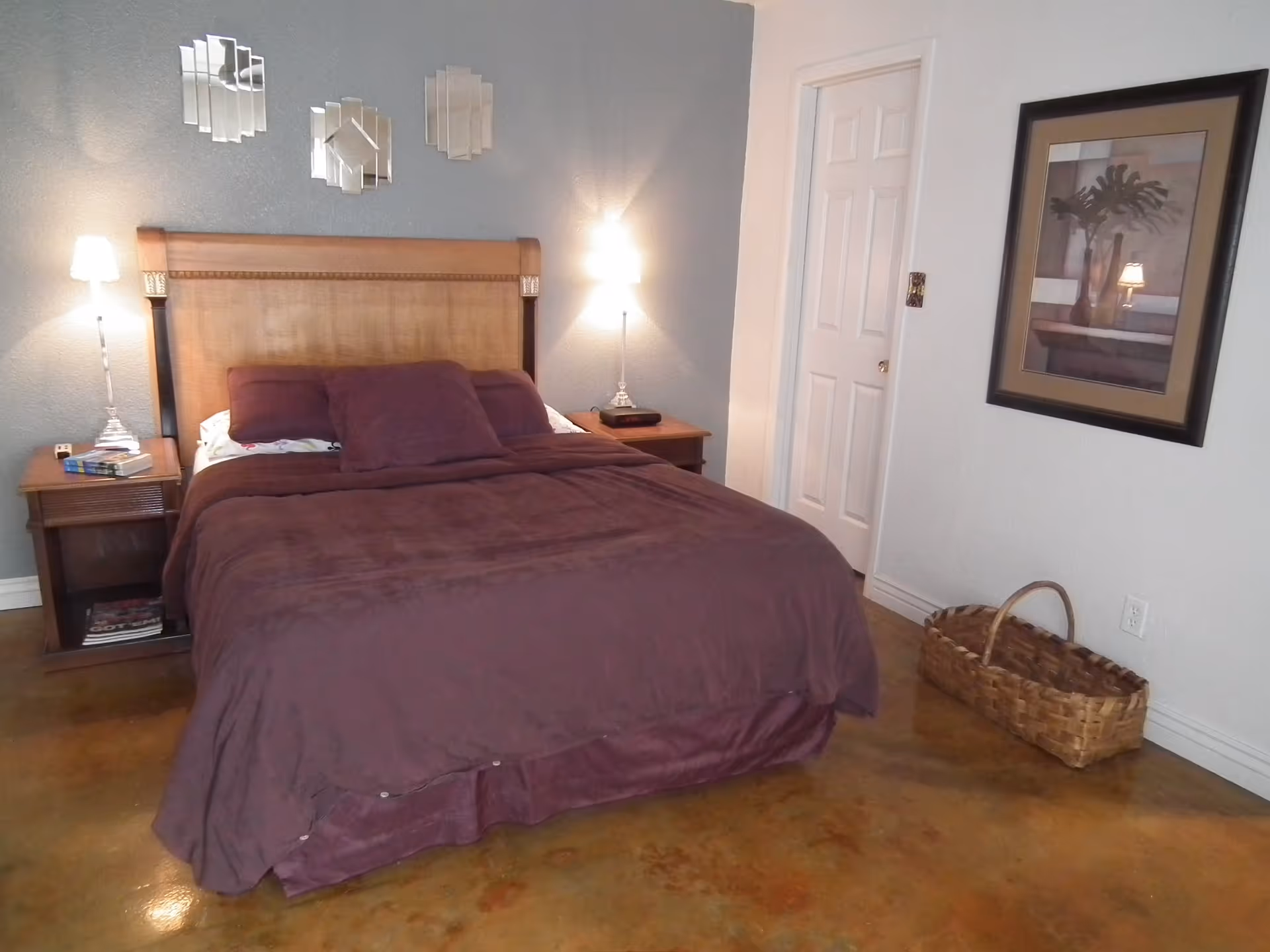 A bedroom with a large wooden headboard bed covered in a dark purple blanket and pillows. There are two bedside tables with lamps on each side of the bed. Above the headboard, three decorative mirrors are mounted on a gray wall. On the right wall, there is a framed picture and a woven basket on the floor. The floor has a polished brown finish.