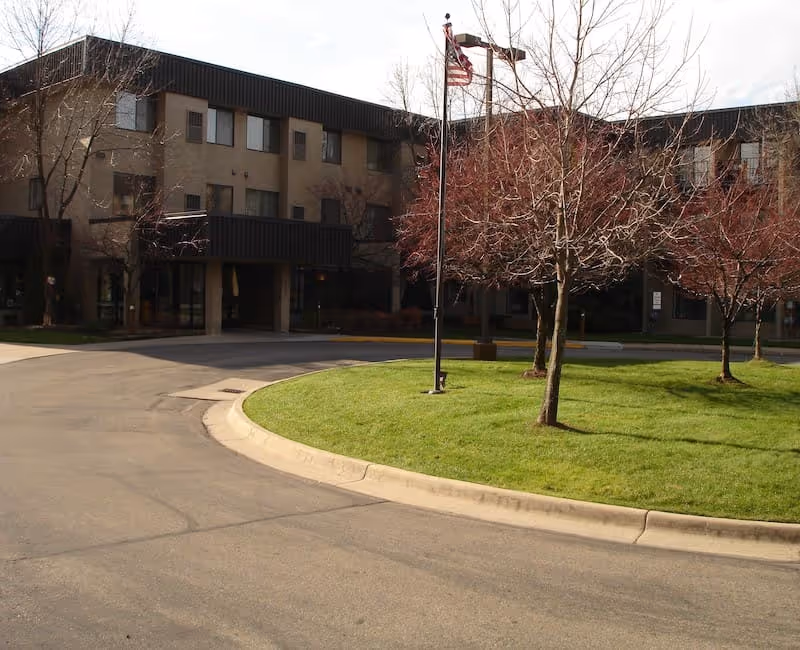Exterior view of Madison Manor Apartments showing a three-story building with multiple windows, a curved driveway, a grassy area with leafless trees, and an American flag on a flagpole.