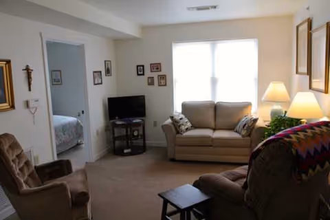 Cozy living room with beige sofa, chairs, TV, lamps, and a doorway to a bedroom.