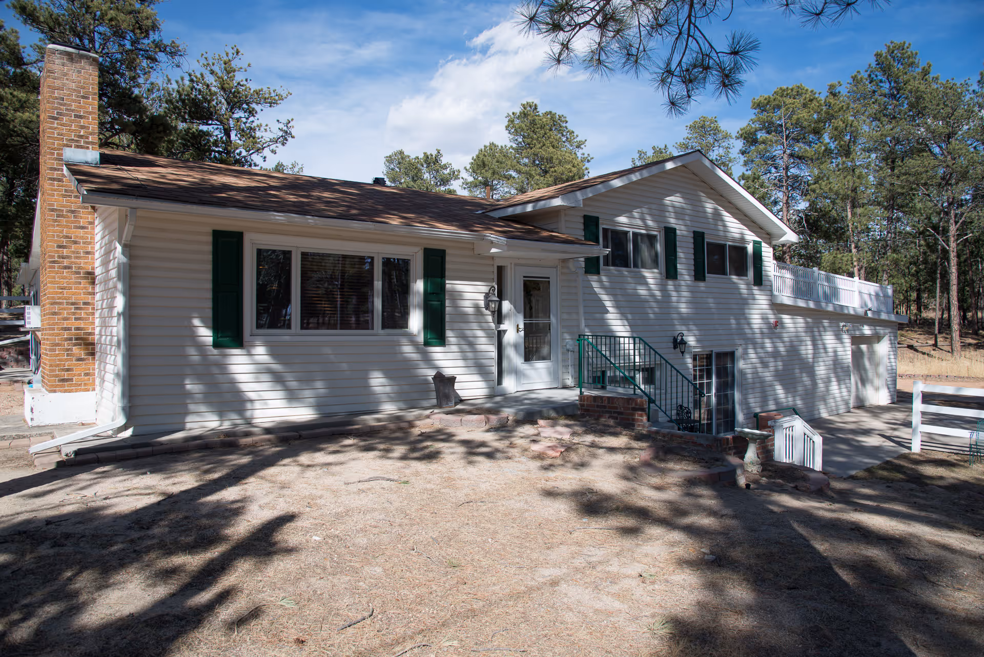 Exterior view of a single-story white house with green shutters and a brown roof, surrounded by trees and a dry yard. The house features a brick chimney, a front door with a small porch and steps, and a garage on the right side.