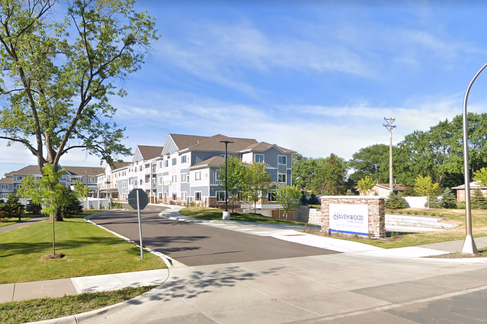 Exterior view of Havenwood of Richfield, a multi-story senior living facility with gray and white siding, surrounded by green trees and a well-maintained lawn under a blue sky.