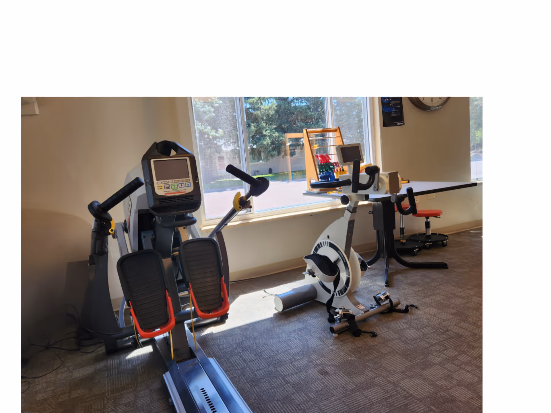 A bright exercise room with stationary and recumbent bikes and other fitness equipment arranged in front of a large window.