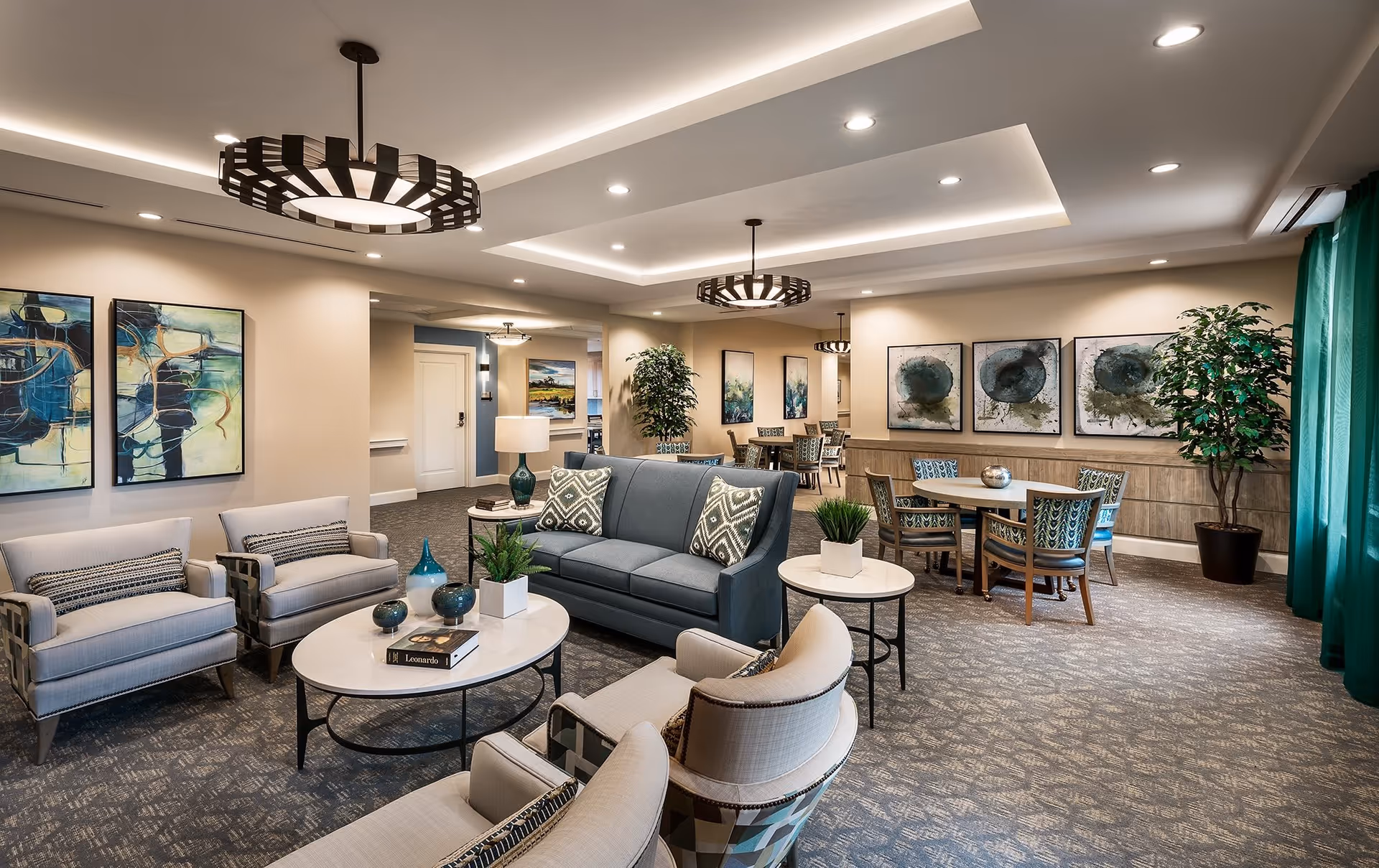 Well-lit communal lounge with sofas, armchairs, coffee tables, dining tables, artwork, and decorative pendant lighting.