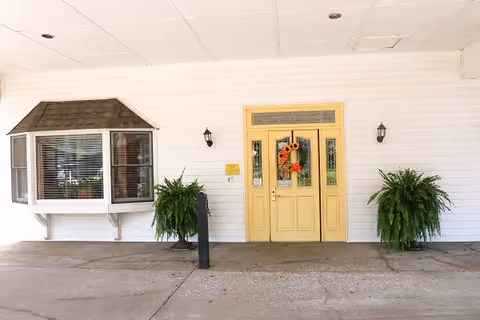 Covered building entrance with a yellow door decorated with a wreath, flanked by potted ferns and a bay window.