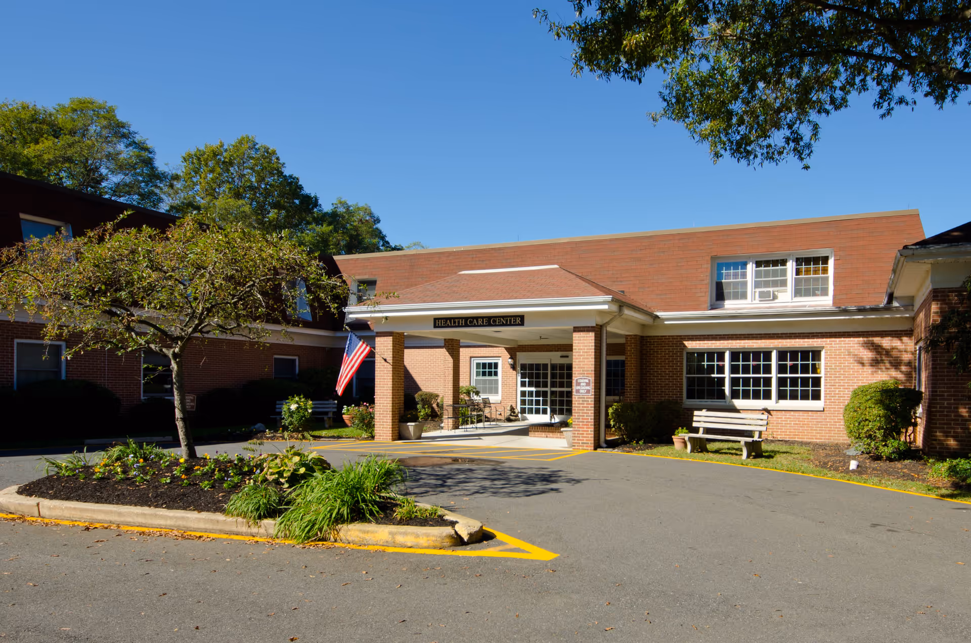 Exterior view of a brick health care center building with a covered entrance, an American flag, a small landscaped area with plants and a tree, and a bench on the right side under a clear blue sky.
