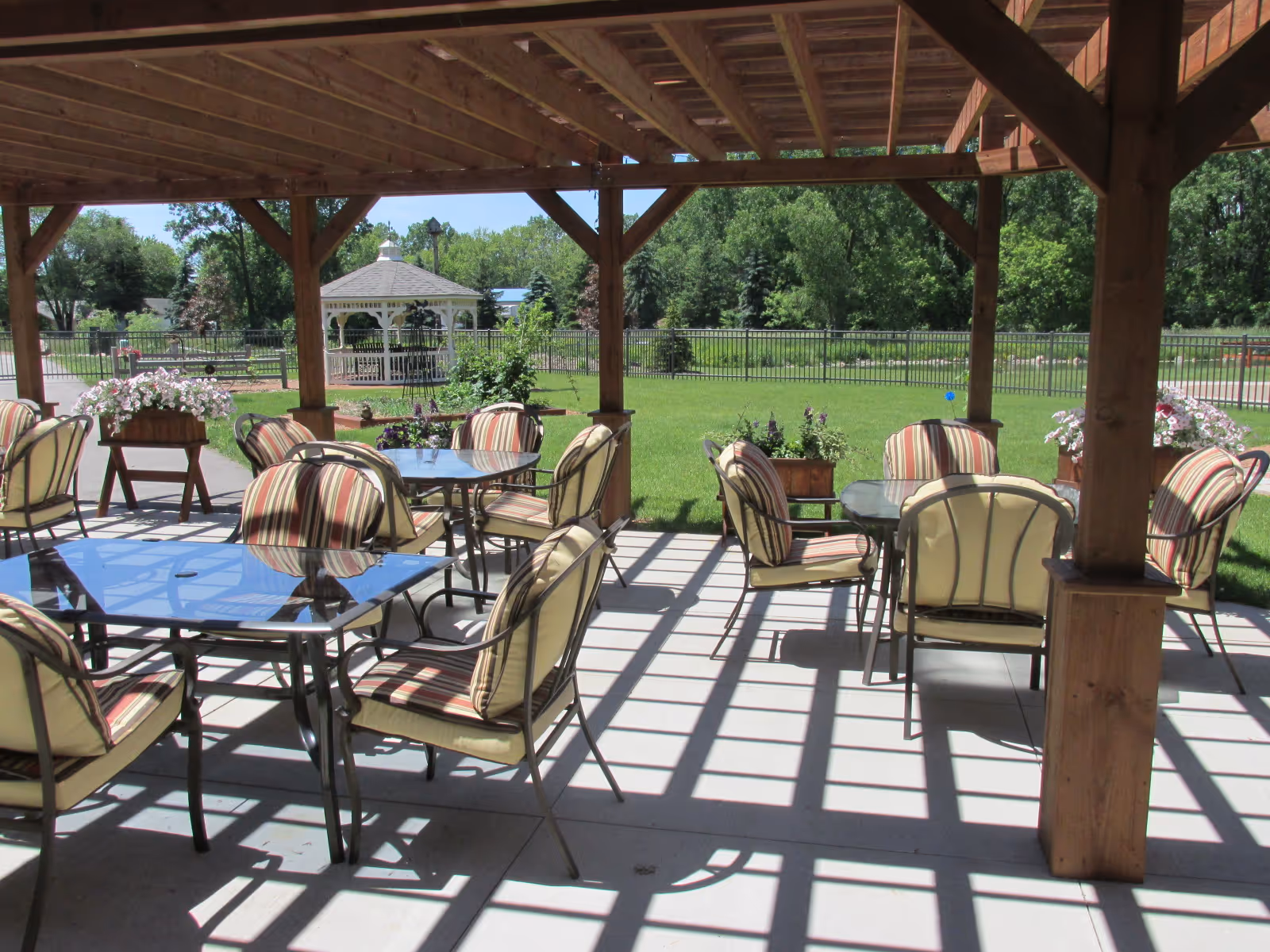Covered outdoor patio with glass-top tables and striped cushioned chairs overlooking a grassy yard and gazebo.