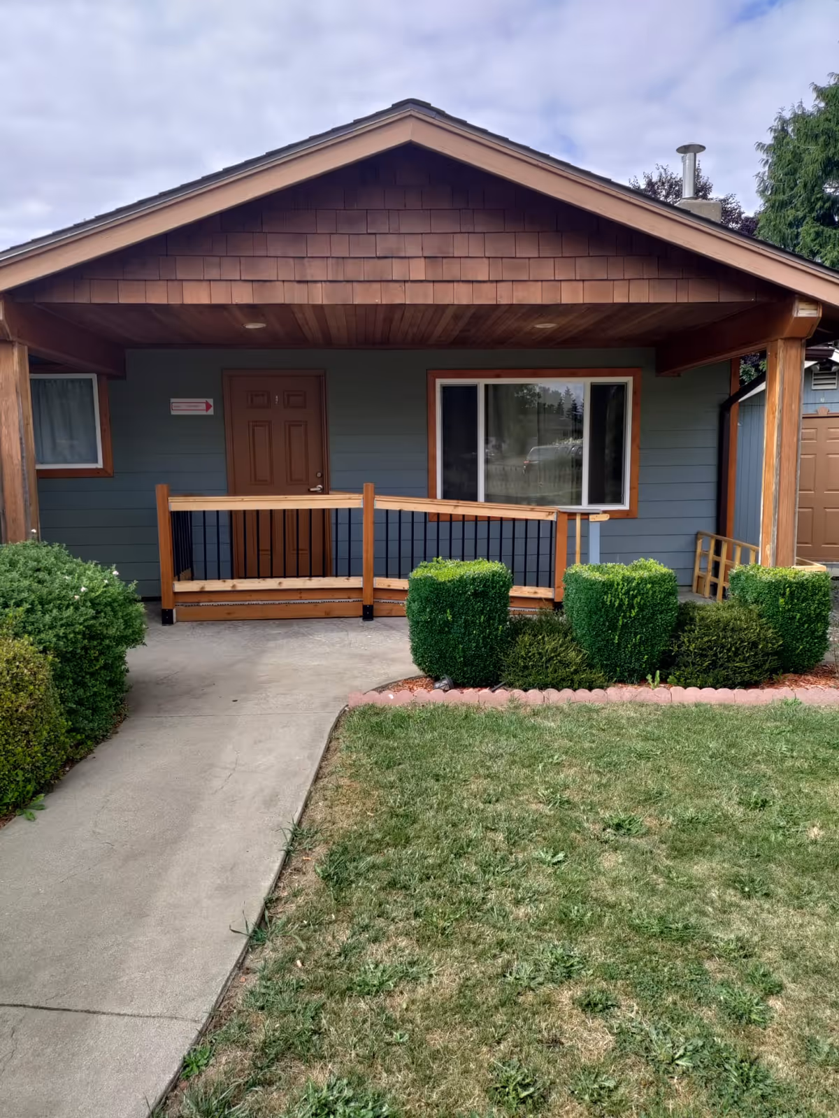 Front porch of a single-story house with a covered entry, wooden railing, brown door, window, concrete walkway and trimmed shrubs.