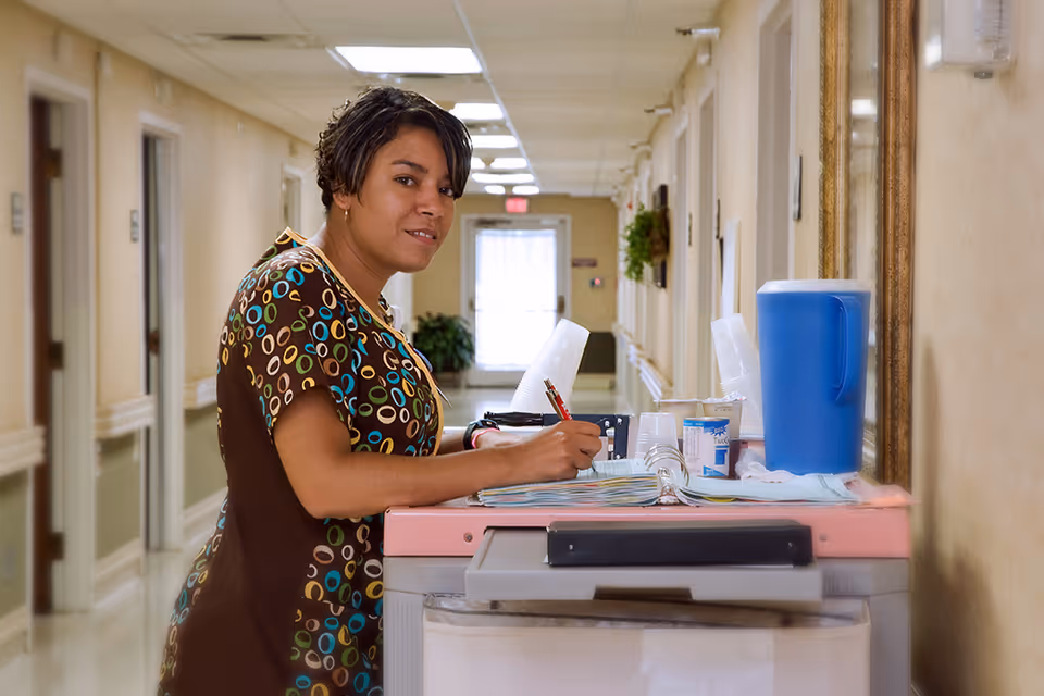 A healthcare worker in a patterned scrub top is standing in a nursing care center hallway, writing in a large open binder on a pink and gray cart that holds a blue pitcher, disposable cups, and other supplies.