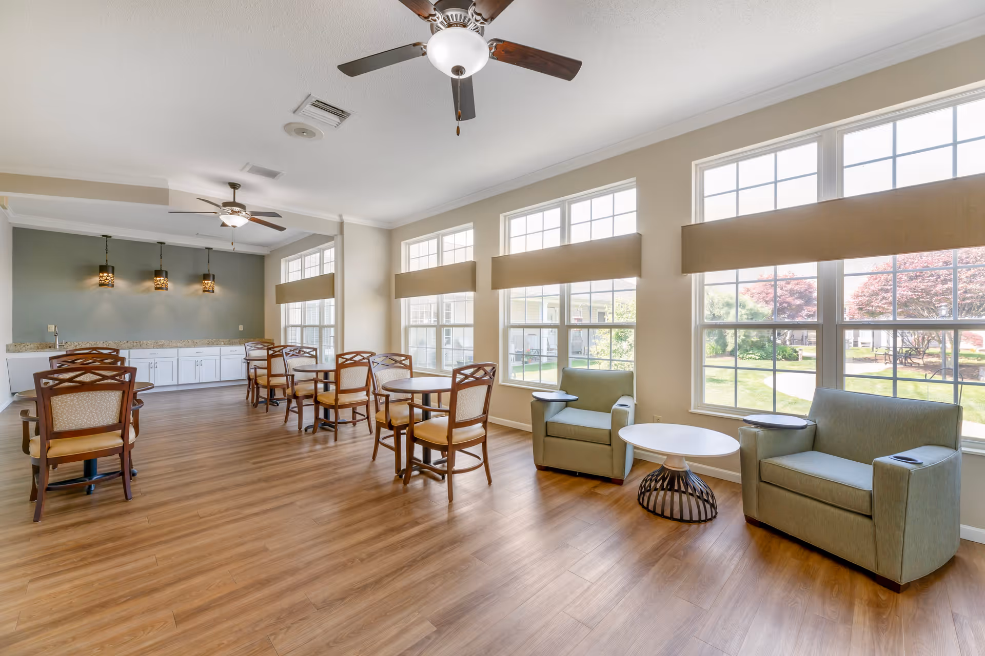 A bright and spacious common area with large windows letting in natural light. The room features wooden flooring, ceiling fans, several round tables with chairs, and two green armchairs with small side tables near the windows. The back wall has white cabinetry and three hanging pendant lights.