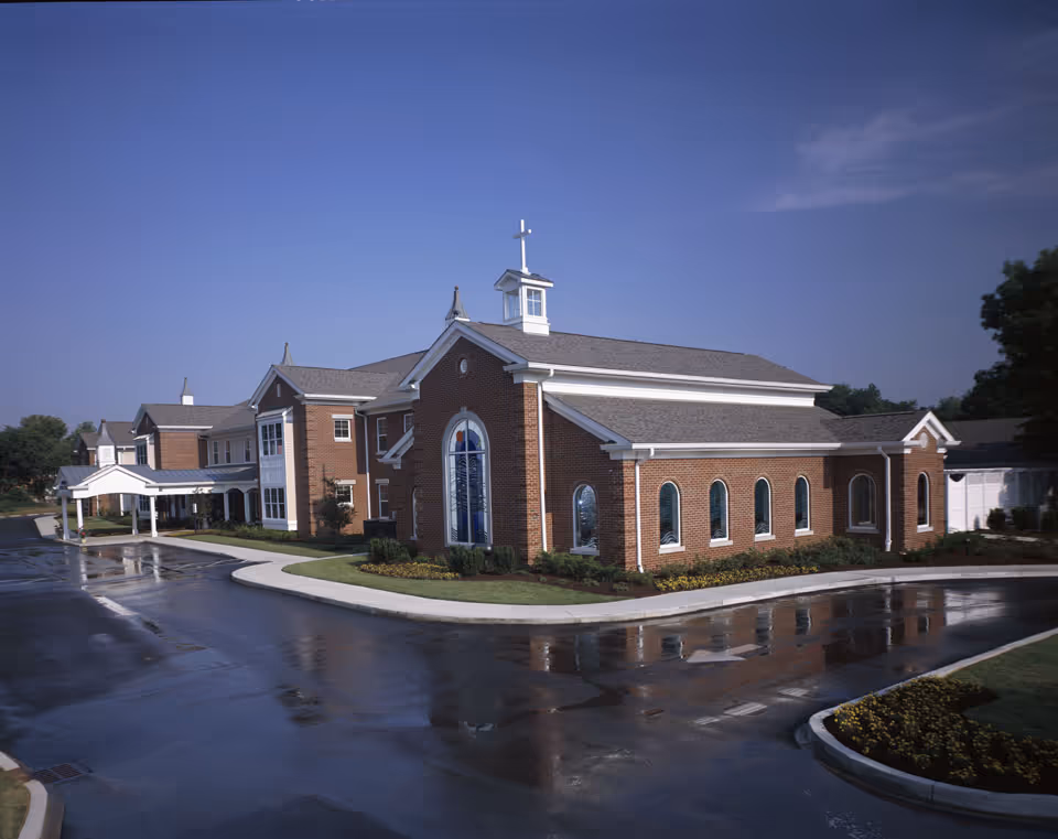 Brick exterior of a senior care building with a chapel-like section topped by a cross and a wet curved driveway in front.