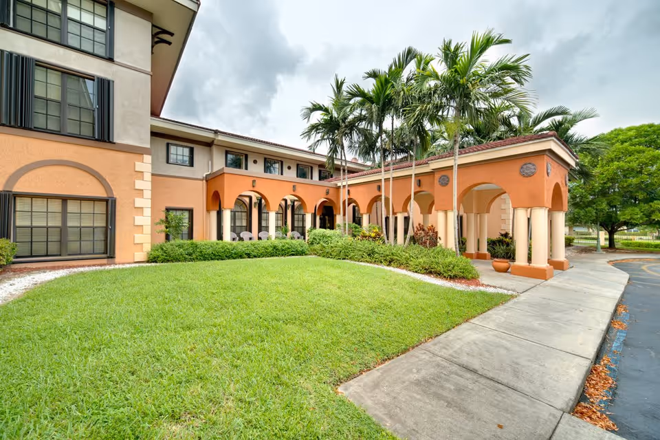 Exterior view of a senior living facility named Presidential Place featuring a two-story building with beige and orange walls, arched walkways, palm trees, green lawn, and a concrete sidewalk leading to the entrance.