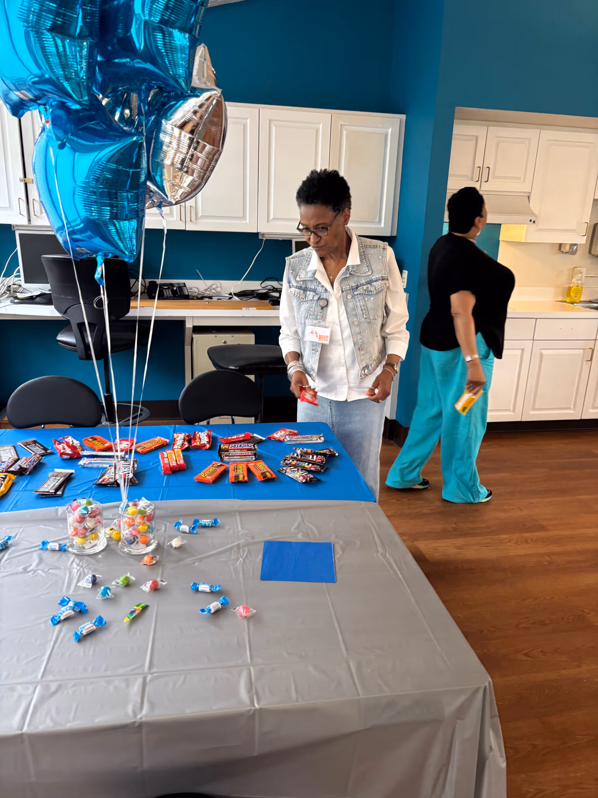 Two women in a room with blue walls and white cabinets. One woman is standing near a table covered with a gray and blue tablecloth, arranging various candy bars and sweets. The other woman is walking away holding a candy bar. There are blue and silver star-shaped balloons tied to the table.