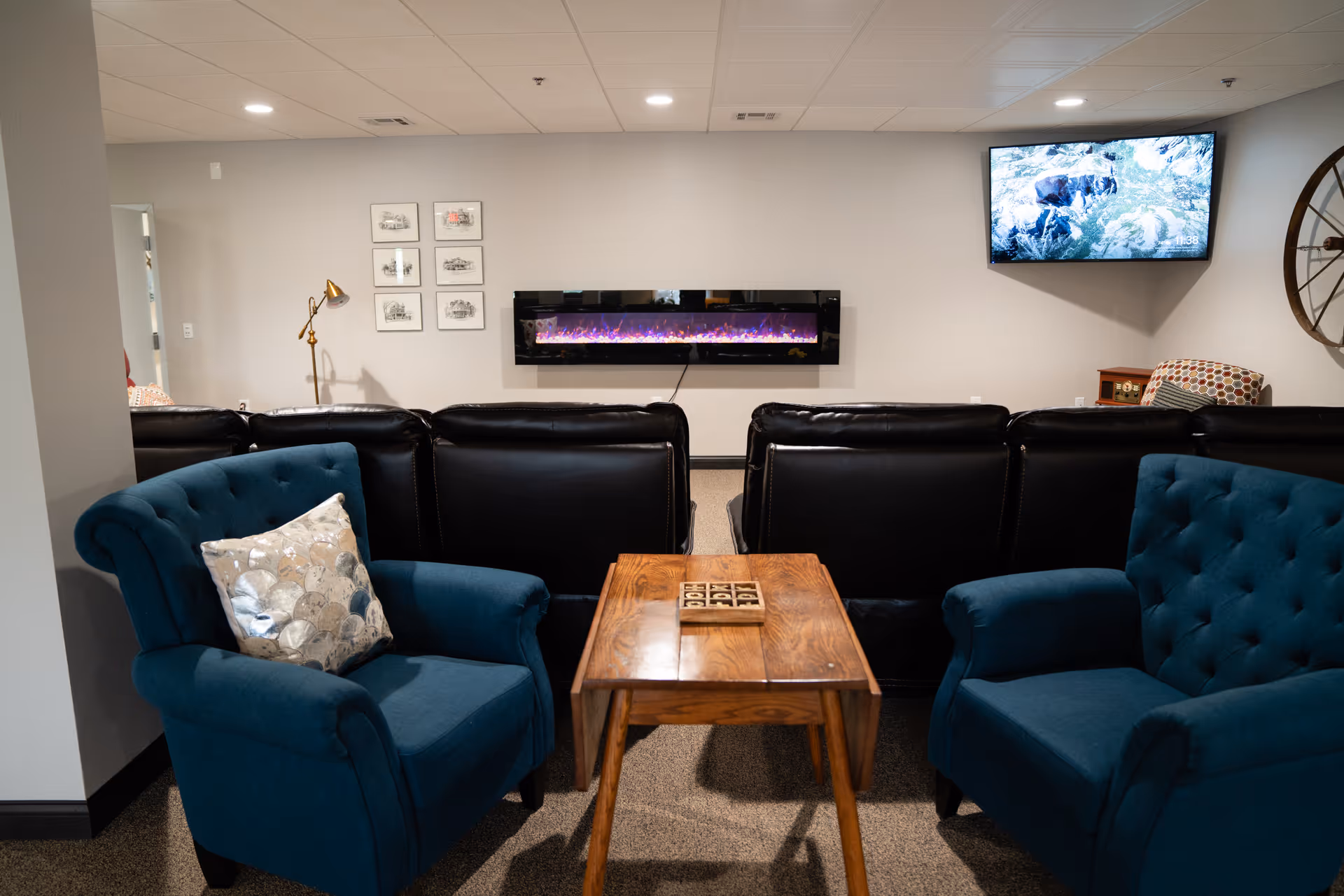 Cozy common living room with teal armchairs, a wooden coffee table, black leather recliners, a wall-mounted electric fireplace and a TV.