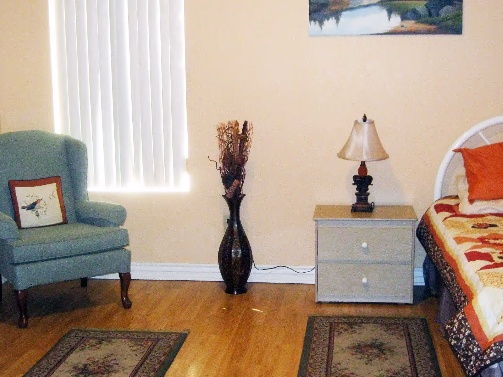 A bedroom with a green armchair, decorative floor vase, a nightstand with a lamp, and a bed topped with a patterned quilt.