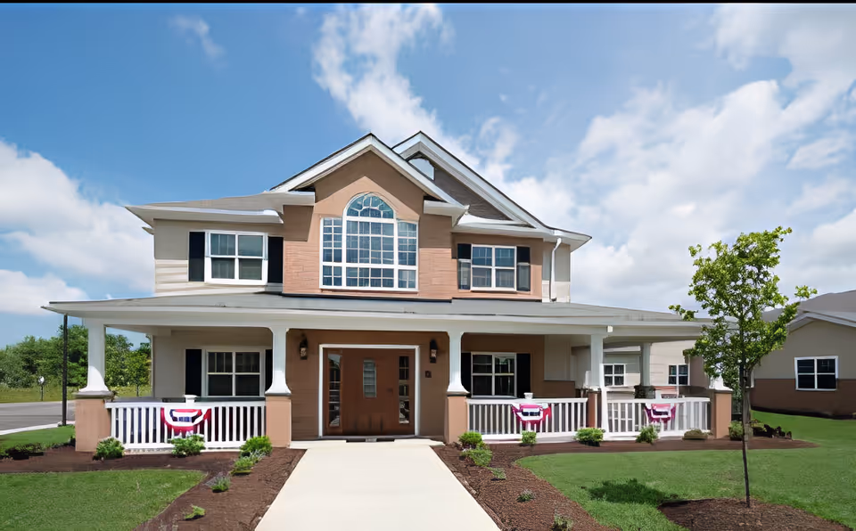 Front exterior view of a two-story residential building with a large porch, white railings decorated with red, white, and blue bunting, and a well-maintained lawn with small shrubs and a young tree under a partly cloudy sky.