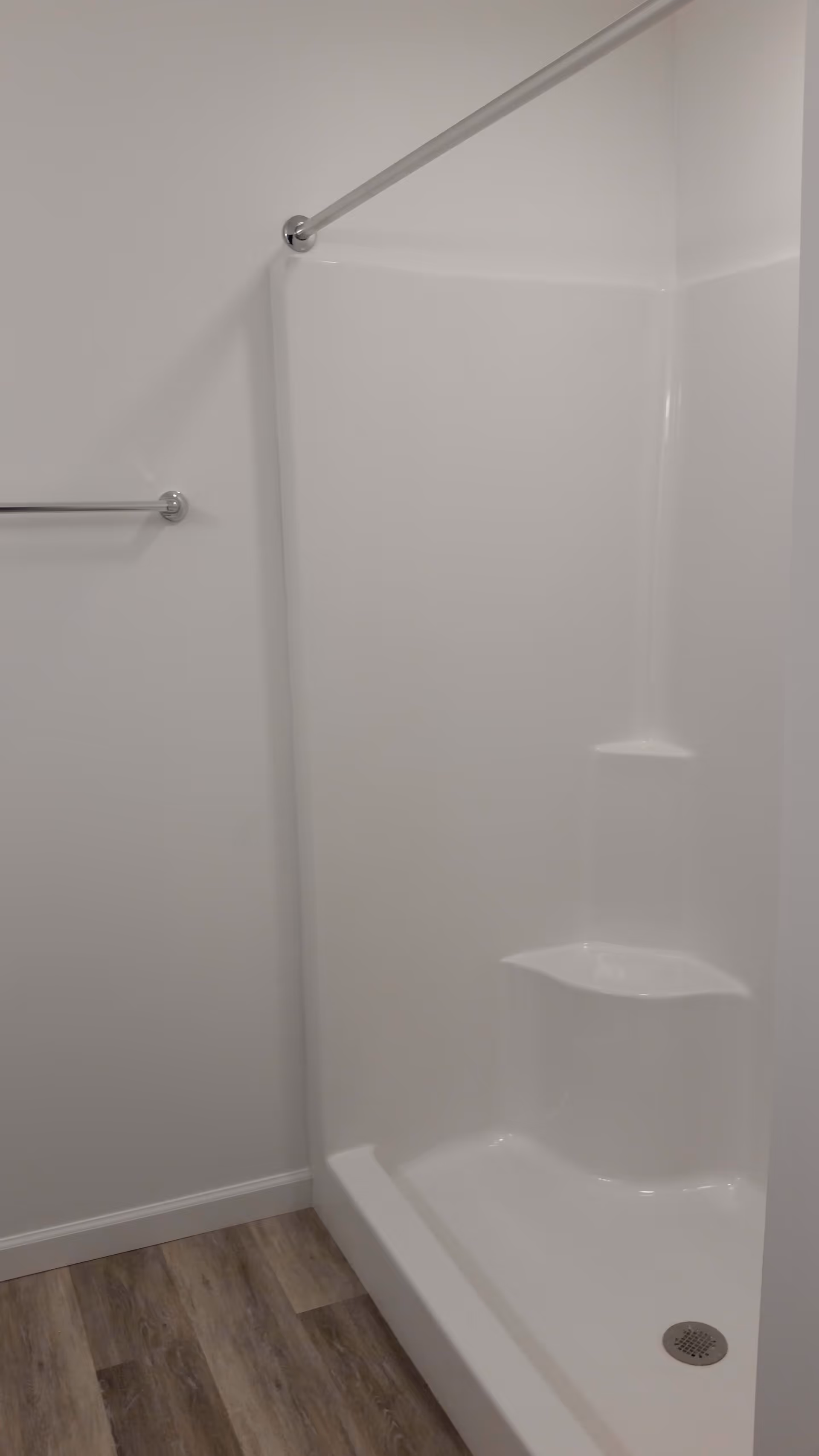 A clean white shower stall with built-in corner shelves and a silver shower curtain rod above. There is a silver towel bar mounted on the adjacent white wall and wood-patterned flooring below.