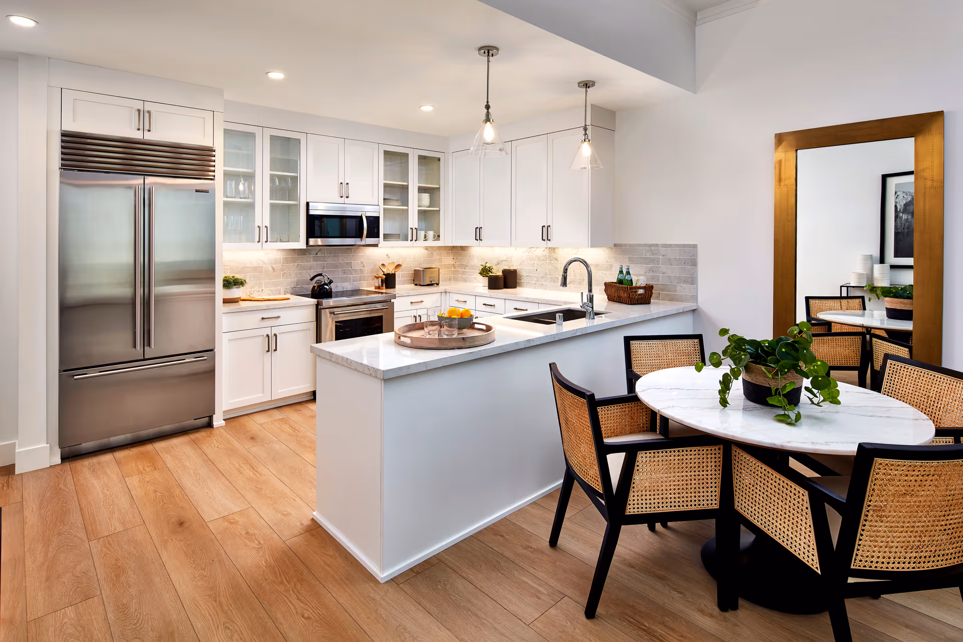 Bright modern kitchen with white cabinetry, a marble island and stainless steel refrigerator, plus an adjacent round dining table with woven chairs and a potted plant.