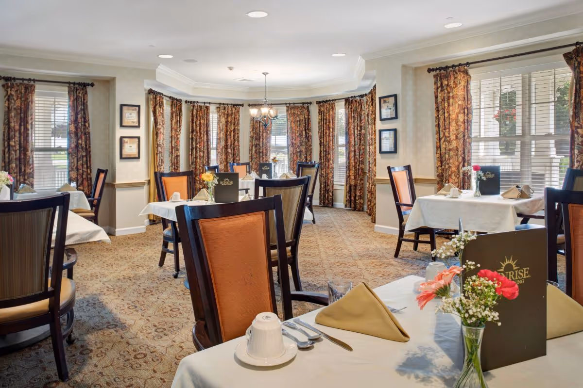 A well-lit dining room with multiple tables covered in white tablecloths, each set with napkins, cups, and silverware. The room features patterned carpet, floral curtains on large windows, and framed certificates on the walls. There are flower vases on the tables and menus labeled 'Sunrise' placed on some tables.