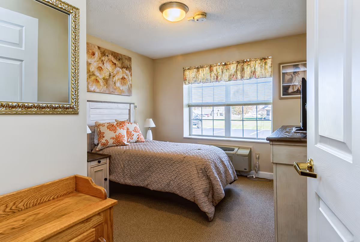 A cozy bedroom with a single bed covered in a beige quilt and two orange floral pillows. The room features a large window with floral curtains, a wooden nightstand with a white lamp, a dresser with a TV on top, a decorative mirror on the wall, and a floral painting above the bed. The carpet is beige and the walls are painted light beige.