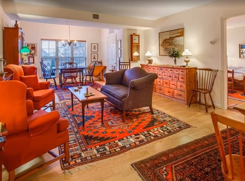 Cozy living room and dining area with red patterned rugs, upholstered chairs and sofa, wooden sideboard and a dining table by a window.