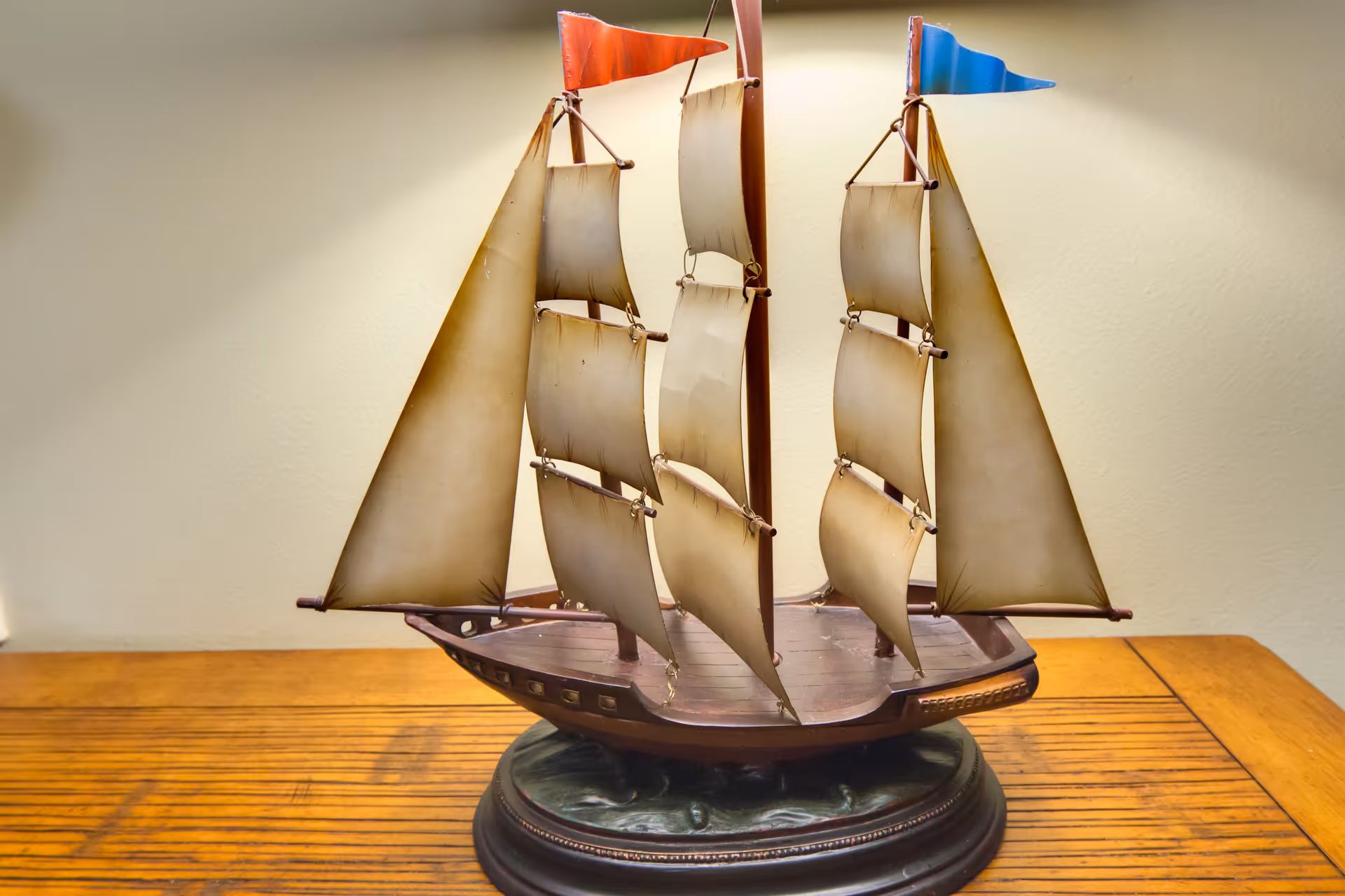 A detailed model of a sailing ship with three masts and beige sails, each topped with a small colored flag, displayed on a wooden surface against a plain light-colored wall.