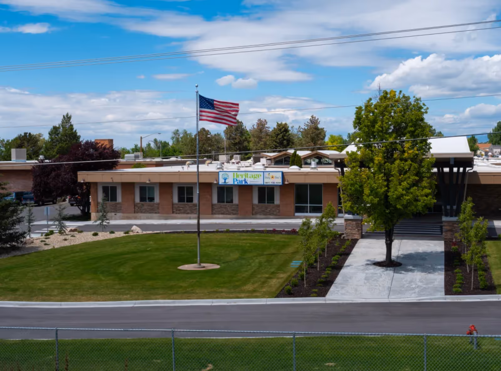 Exterior view of Heritage Park Healthcare and Rehabilitation building with a well-maintained lawn, an American flag on a flagpole, trees, and a clear blue sky with some clouds.
