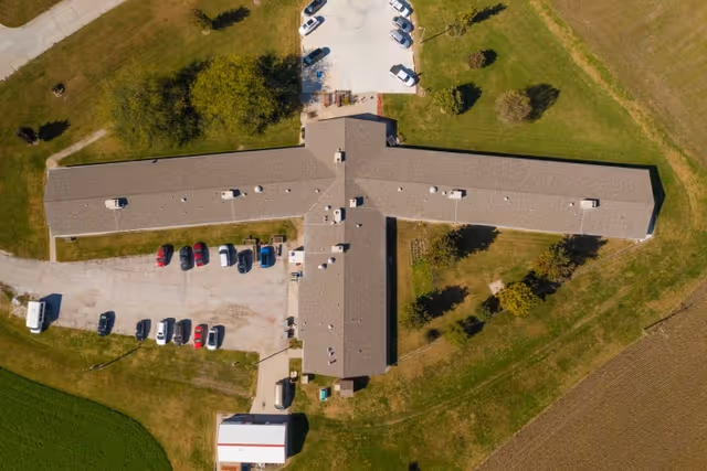 Aerial view of a large, T-shaped building with a parking lot containing several cars. The building is surrounded by green grass and trees, with a small structure nearby and open fields around the property.