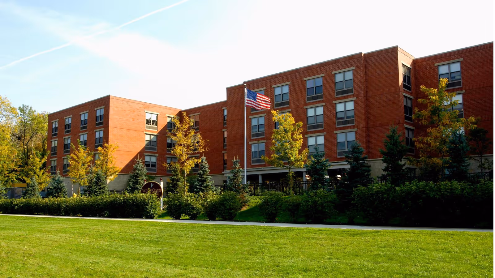 Exterior view of a multi-story brick building with numerous windows, surrounded by trees and shrubs, with a well-maintained green lawn in the foreground and an American flag flying on a flagpole near the entrance.