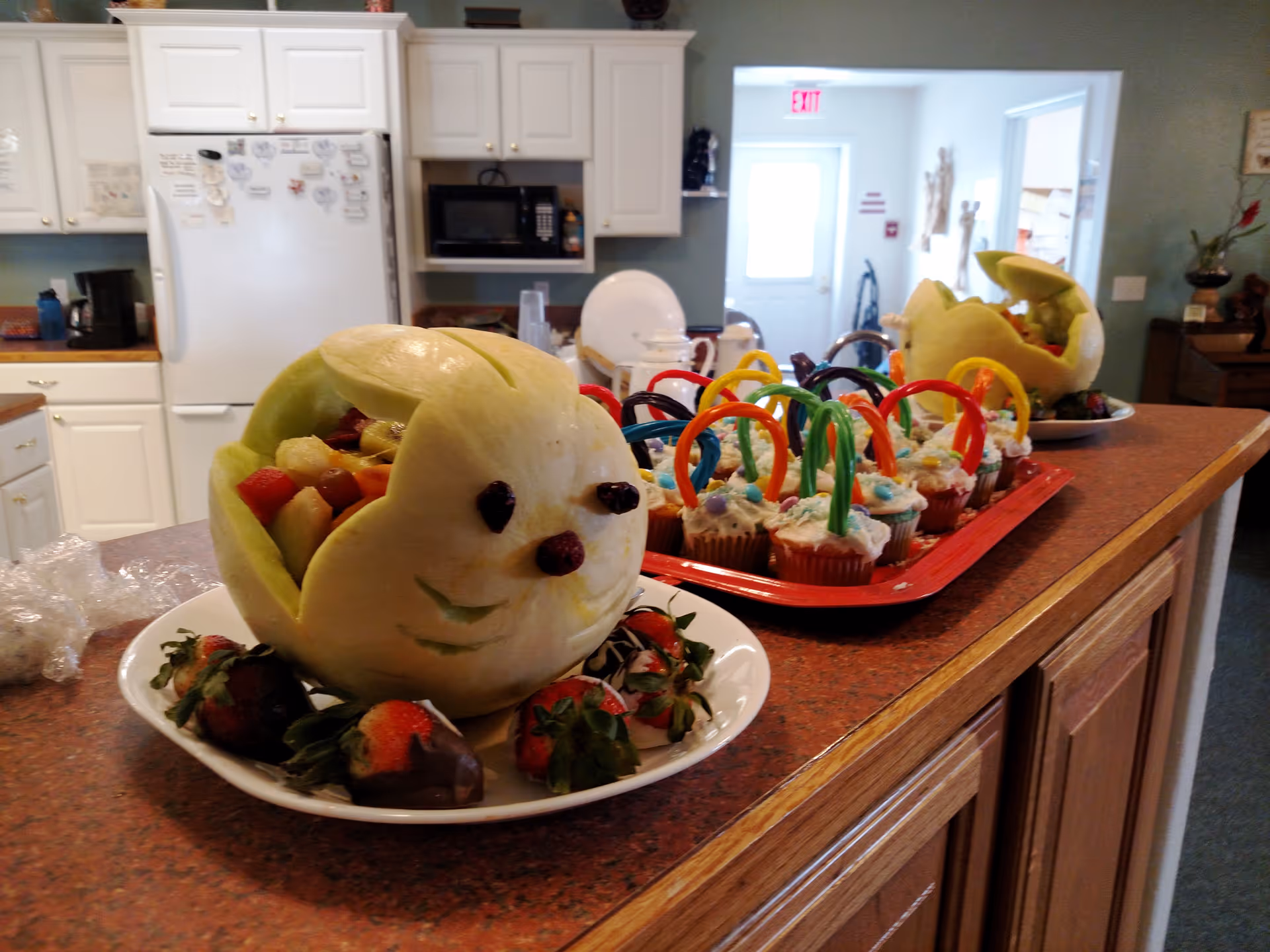 A kitchen counter with a carved melon shaped like a bunny filled with fruit, surrounded by chocolate-covered strawberries on a white plate. Behind it, there is a tray of cupcakes decorated with colorful candy handles resembling baskets. The kitchen has white cabinets, a refrigerator with magnets, and a microwave. The background shows a hallway with a door and some wall decorations.