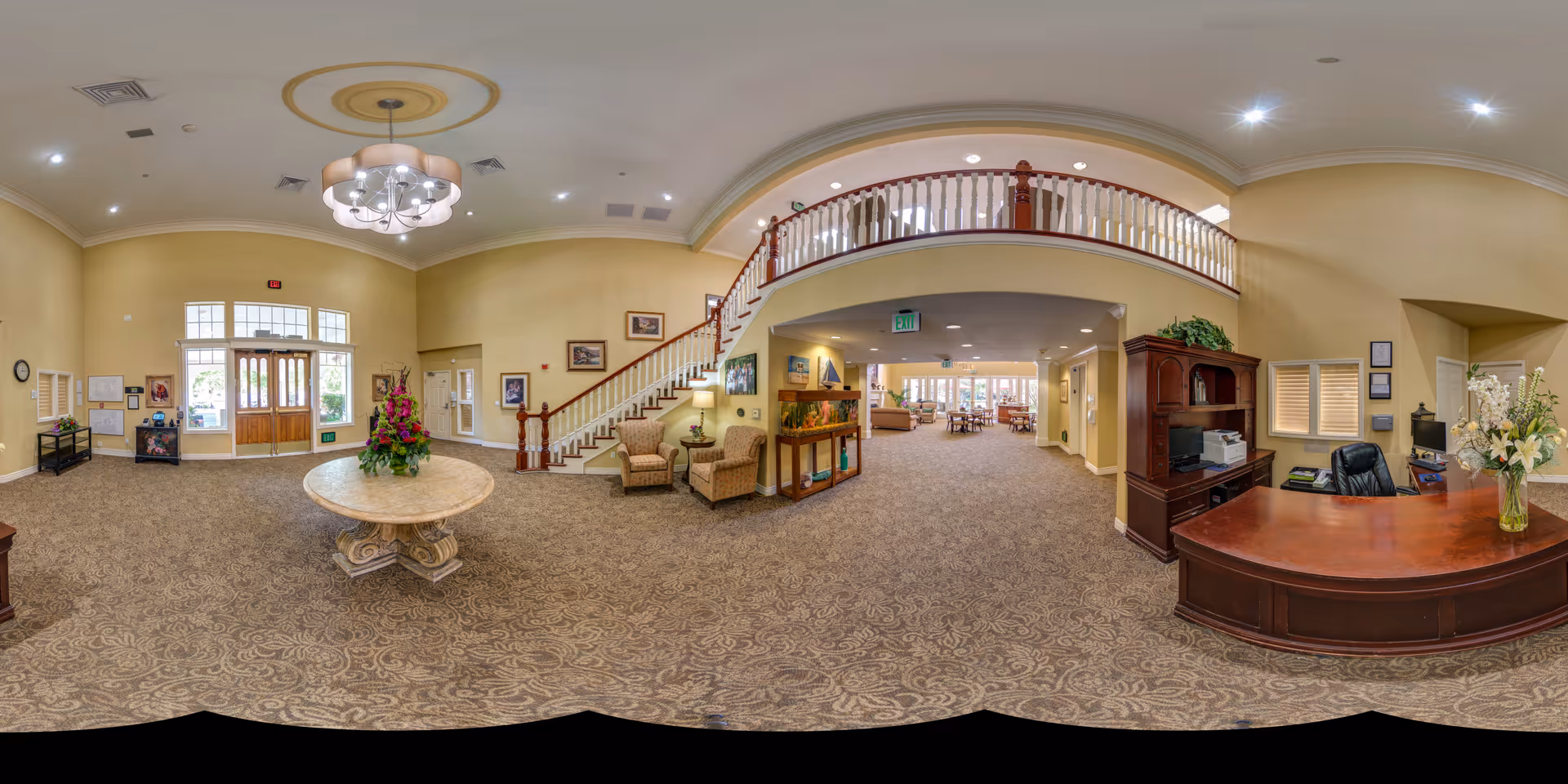 A spacious and well-lit senior living facility lobby with beige walls and patterned carpet. The room features a round table with a floral arrangement in the center, a wooden reception desk with a vase of flowers, comfortable seating areas with armchairs, a staircase with wooden railing leading to an upper floor, and large windows and doors letting in natural light.