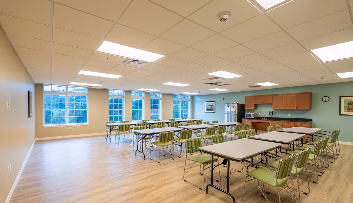 A spacious, well-lit room with multiple rectangular tables and green striped chairs arranged in rows. The room has large windows on one side letting in natural light, light wood flooring, and a kitchenette area with wooden cabinets, a refrigerator, microwave, and sink against a green wall. There are framed pictures on the walls and a clock above the kitchenette.