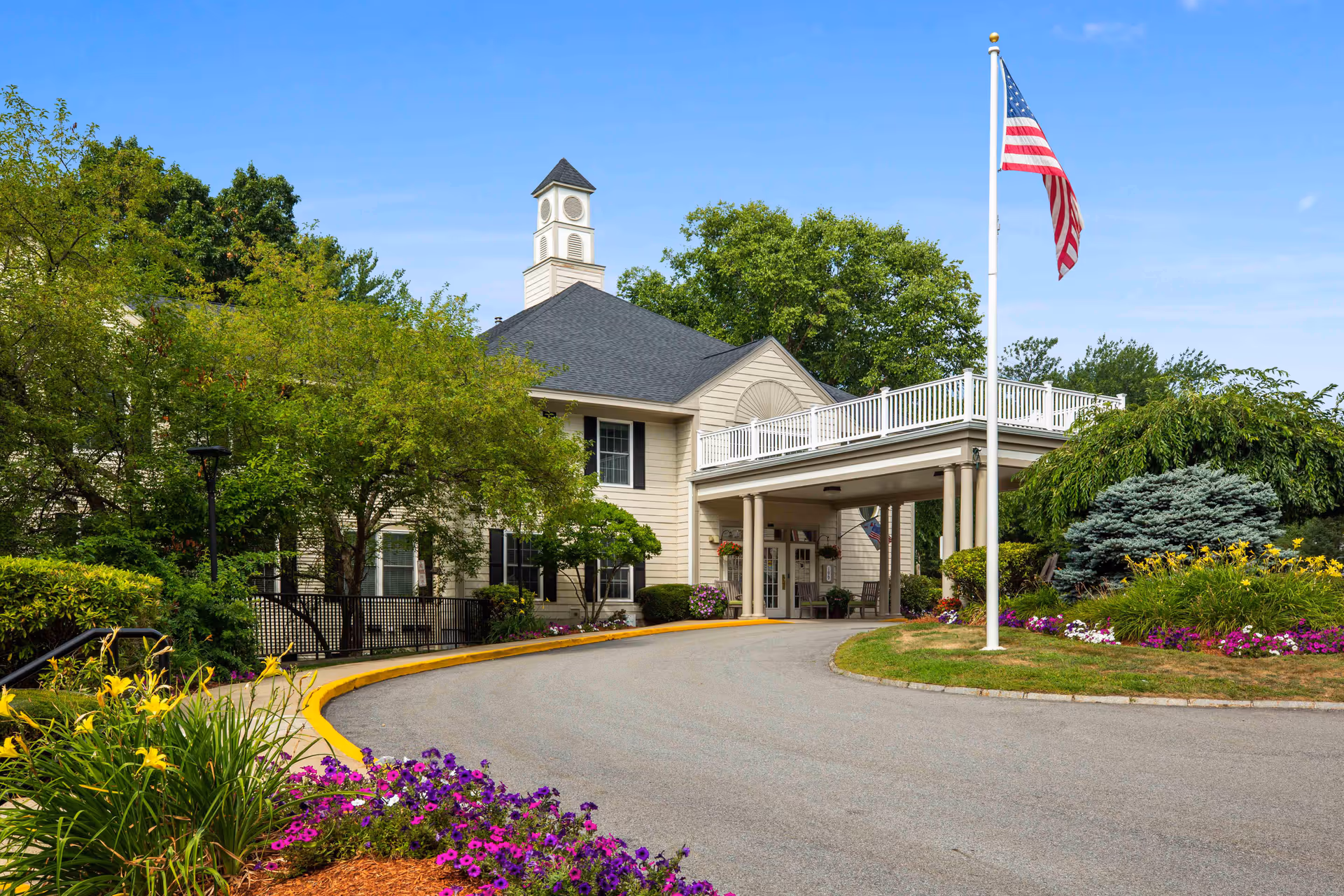 Exterior view of a senior living facility with a driveway leading to a covered entrance. The building has beige siding, black shutters, and a small cupola on the roof. There is a tall flagpole with an American flag, surrounded by well-maintained landscaping including colorful flowers, bushes, and trees under a clear blue sky.