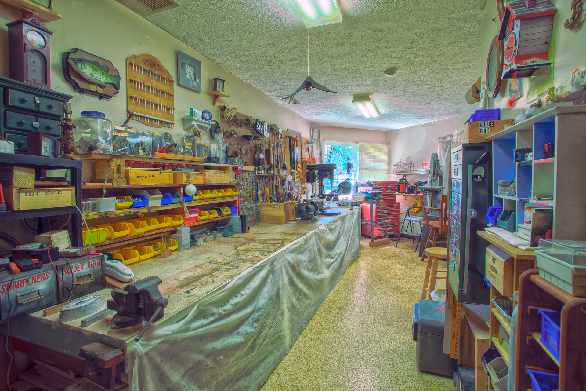 A well-organized workshop room with a long workbench covered by a cloth, various tools hanging on the walls, shelves filled with small bins and containers, and equipment such as a drill press and vise. The room is lit by fluorescent ceiling lights and has a window at the far end letting in natural light.