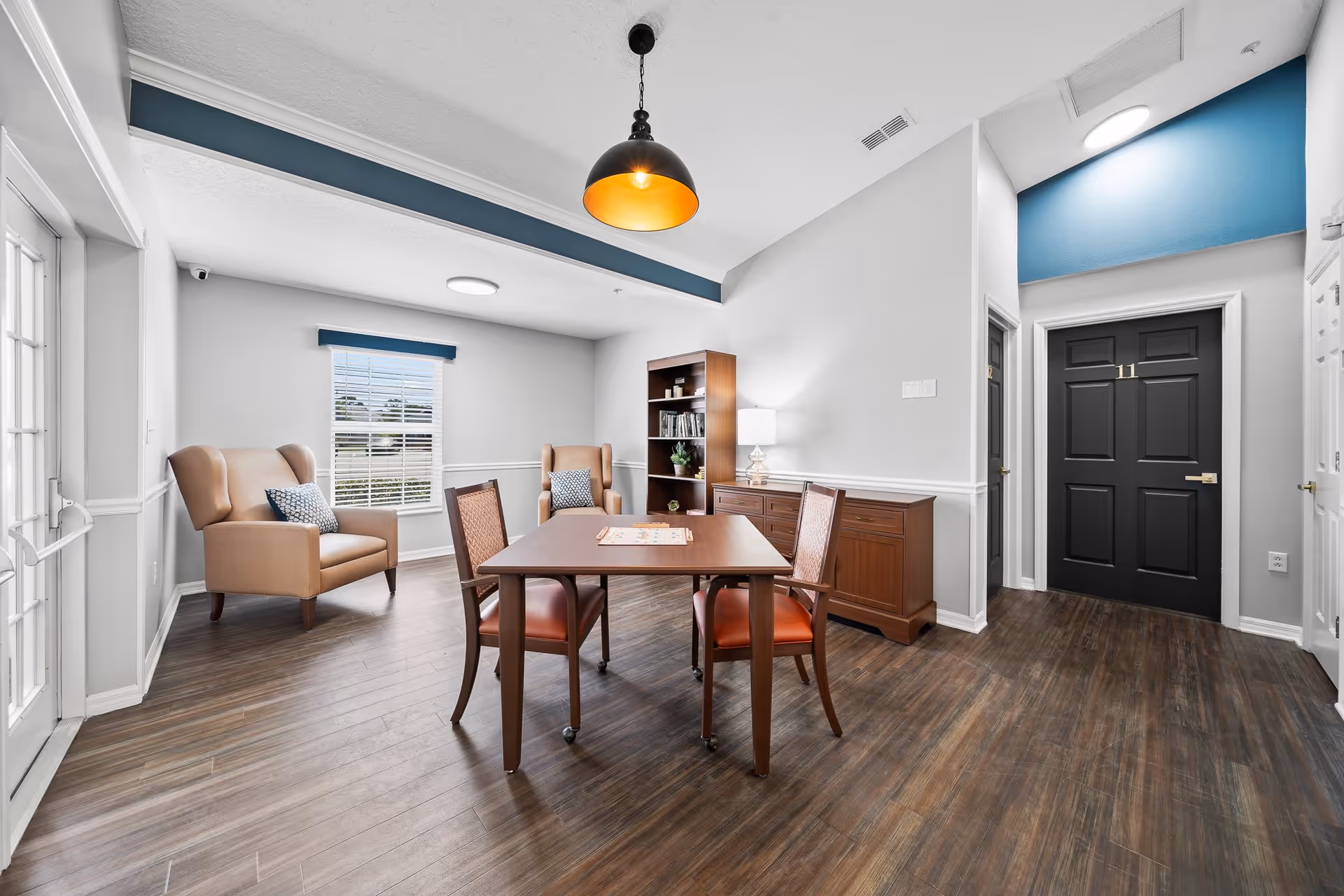 A bright and clean common area in a senior living facility featuring a wooden table with four chairs, two beige armchairs with patterned cushions near a window, a wooden bookshelf, and a sideboard with a lamp. The room has wood flooring, white walls with blue accents, and a black door numbered 11.