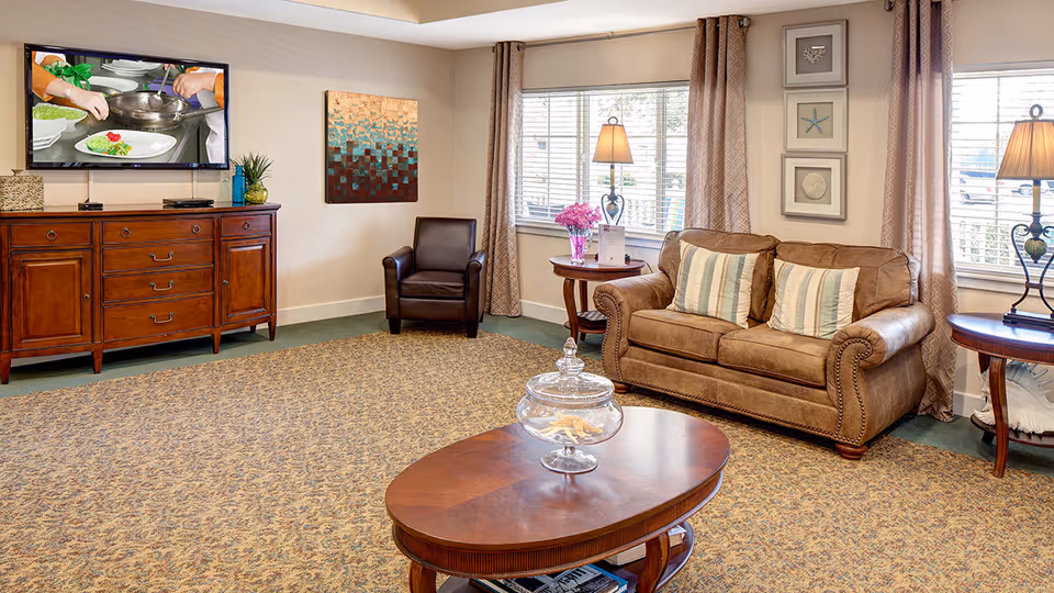 Bright communal living room with a leather sofa and armchair, coffee table, TV on a wooden console, and windows with lamps.