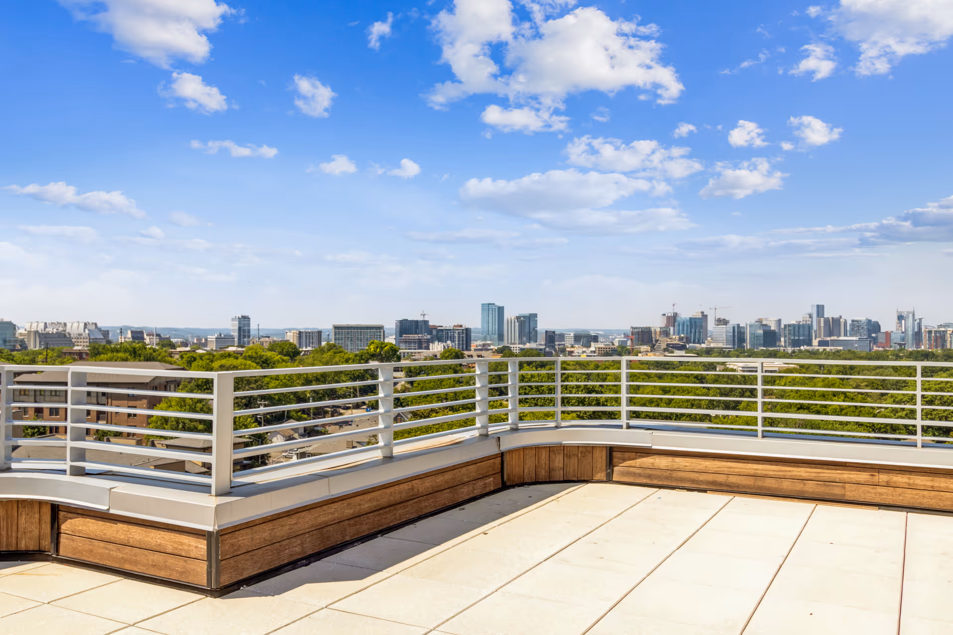 View from a rooftop terrace with a white metal railing and wooden paneling, overlooking a city skyline under a blue sky with scattered clouds.