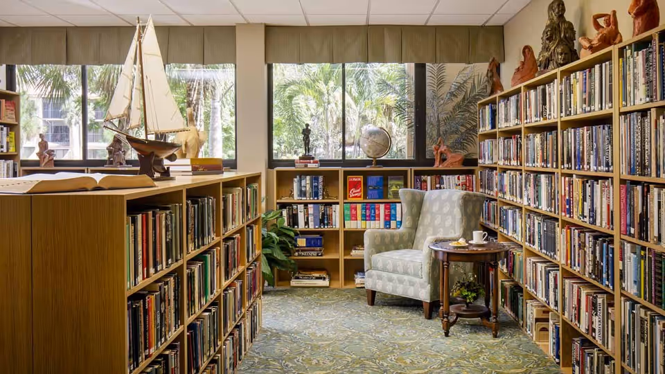 Cozy interior library with wooden bookshelves, an upholstered armchair and side table, and large windows showing palm trees outside.